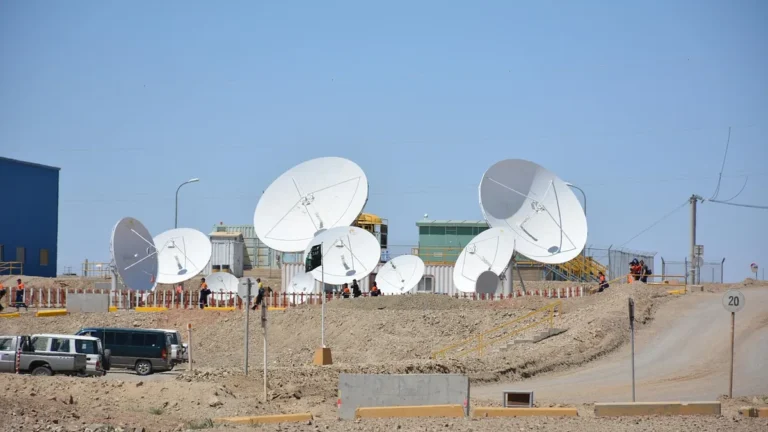 Large white satellite dishes on a dusty hillside with workers and vehicles under a clear blue sky.