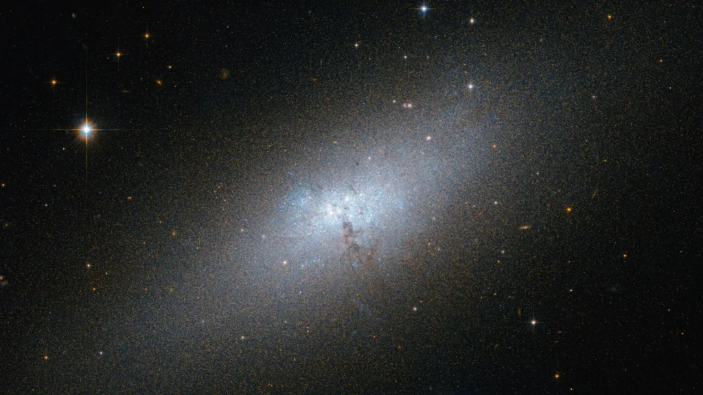 A luminous blue dwarf galaxy glows in a vast field of stars, with a bright star shining on the left.