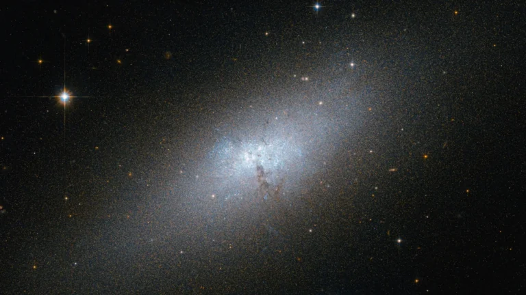 A luminous blue dwarf galaxy glows in a vast field of stars, with a bright star shining on the left.