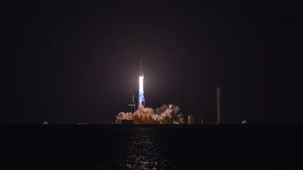 A rocket launches into the night sky, with bright fire and billowing smoke reflecting on the water below.