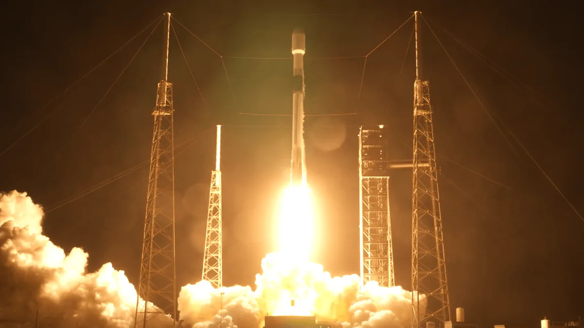 A rocket launches into the night, glowing brightly with fire and smoke around metal towers.