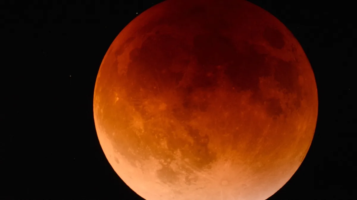 A close-up of a glowing orange-red moon showing surface craters during a total lunar eclipse against a black sky.
