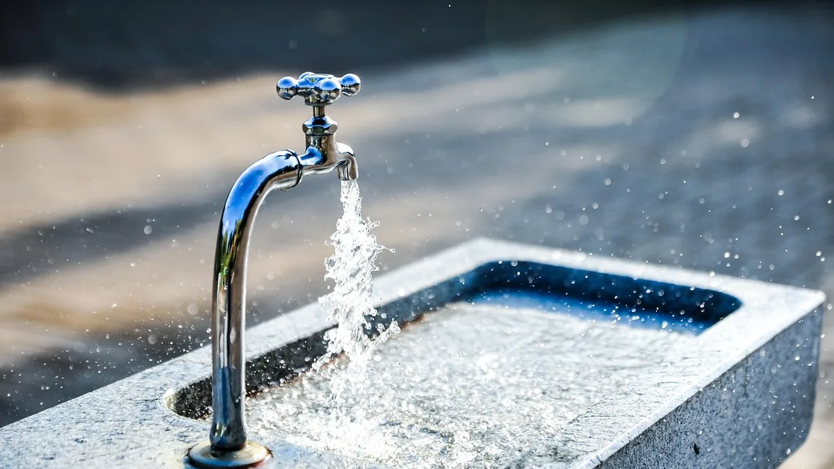 A shiny metal faucet pours splashing water into a stone basin under bright sunlight.