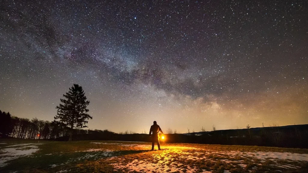 Person silhouetted holding a bright light under a star-filled Milky Way sky over a snowy field.