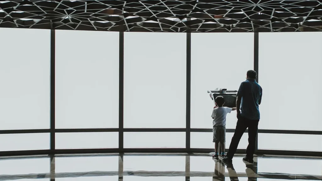 Silhouette of a father and son looking through an observation telescope against a bright, large window under a geometric
